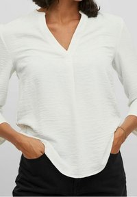 Blouse blanche texturée avec un col en V et des manches trois-quarts. Présente une coupe décontractée et un ourlet arrondi. Portée avec les mains dans les poches.