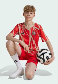 Equipación de fútbol roja que presenta una camiseta con un patrón y acentos dorados, junto con pantalones cortos a juego. Botas blancas y un balón de fútbol en blanco y negro incluidos.