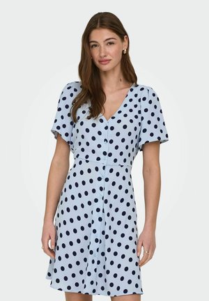 Kvinde med langt brunt hår iført en lyseblå kjole med sorte prikker og korte ærmer, stående foran en ensfarvet baggrund.