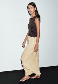 Top senza maniche marrone abbinato a una gonna a più strati di un delicato giallo. L'outfit è completato da sandali piatti marroni e accessori minimali.