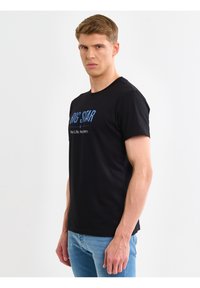 Mladić s kratkom kosom, nosi crnu majicu s natpisom "BIG STAR" i svijetloplave traperice, stoji na jednobojnoj bijeloj pozadini.