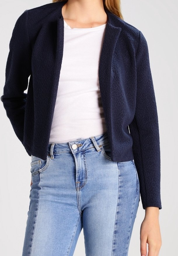 Blazer texturé bleu marine avec une coupe ajustée, fermeture avant simple et manches longues, associé à un haut blanc et un jean bleu clair.