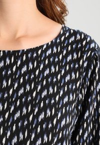 Černý top ze širokého materiálu s volným střihem, zdobený modro-bílým abstraktním vzorem. Výstřih do tvaru lodičky a jemná textura.