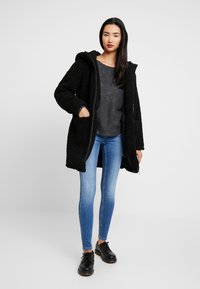 Manteau noir texturé à capuche avec fermeture éclair à l'avant et poches latérales, porté sur un pull gris et un jeans skinny bleu. Des chaussures noires complètent le look.