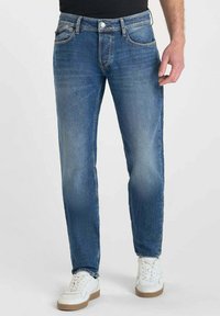 Hombre vestido con jeans ajustados azules y zapatillas blancas, de pie contra un fondo claro y sencillo.