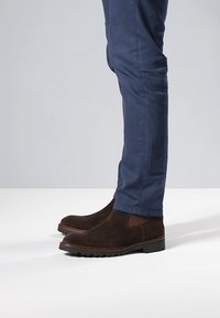 Braune Wildleder-Stiefeletten mit elastischen Seitenpaneelen, Gummisohlen und rundem Zehenbereich, kombiniert mit eng anliegenden marineblauen Hosen.