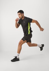 Czarna koszulka sportowa z limonkowymi akcentami i logo, w połączeniu z czarnymi szortami i sportowymi czarnymi butami, model w ruchu, w pomieszczeniu.