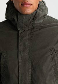 Hombre con chaqueta verde oscuro con capucha, cuello alto, cremallera frontal y bolsillos laterales, sobre un fondo liso.