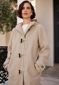 Beige dufflecoat med hætte, lavet af blødt uld, med toggles lukninger, to sidelommer og syet detaljer.