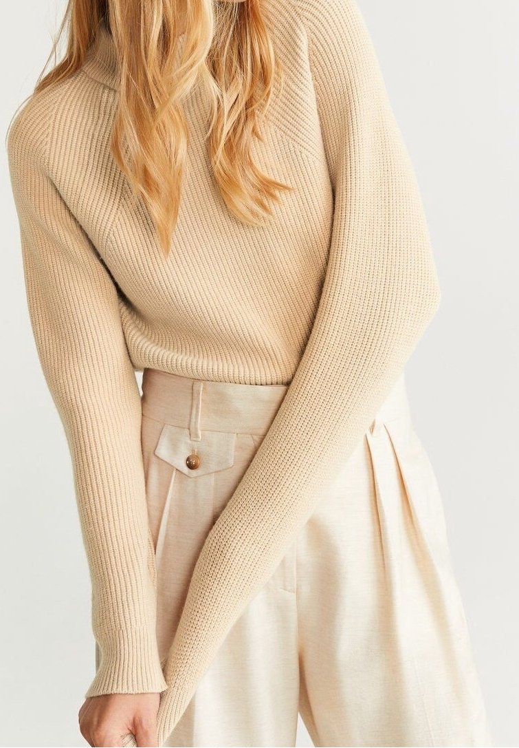 Femme portant un pull côtelé beige rentré dans un pantalon crème taille haute avec un rabat de poche boutonné, posant bras croisés.