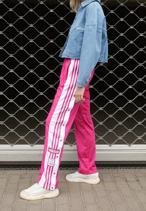 Person iført lyserøde Adidas træningsbukser med hvide striber og hvide sneakers, stående foran en metalport.