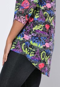 Seitenansicht einer Person, die ein Shirt mit Graffiti-Druck und bunten Wörtern und Formen trägt, kombiniert mit dunklen Hosen vor einem schlichten Hintergrund.