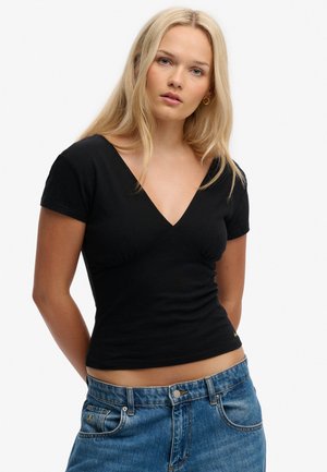 Ung kvinde med langt blondt hår iført en sort V-hals kortærmet top og blå jeans, der vender frem mod en hvid baggrund.