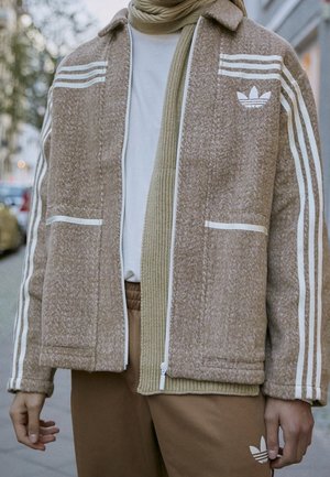 Persona con giacca beige Adidas con strisce bianche, sciarpa a maglia beige, camicia bianca e pantaloni Adidas marroni in piedi all'aperto.
