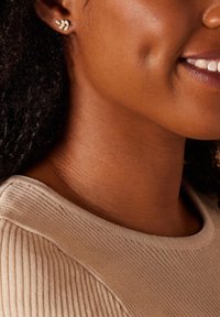 Goldblatt-förmige Ohrstecker werden getragen, sie ergänzen die lockigen Haare und einen beigen Rippensweater, wodurch die Textur und der Farbkontrast hervorgehoben werden.