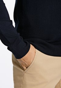 Granatowy sweter dziergany z prążkowanymi mankietami i krawędzią, noszony na beżowych spodniach, z ręką w kieszeni. Gładka faktura, dopasowany krój.