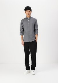 Camisa gris de botones con cuello, mangas enrolladas, combinada con pantalones negros y zapatillas blancas, sobre un fondo liso.