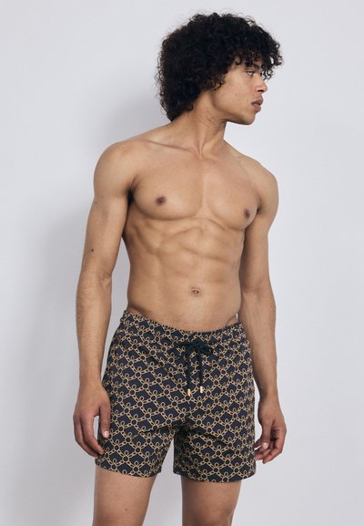 Uomo a torso nudo con capelli ricci, che indossa pantaloni da bagno neri con un motivo a catena dorata, che guarda verso destra su uno sfondo semplice.