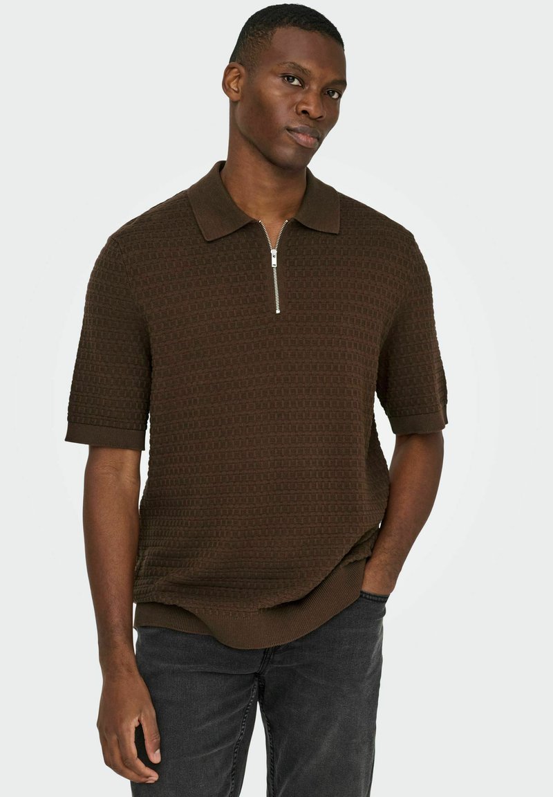Polo-shirt en maille marron avec col zippé, manches courtes, motif texturé et poignets côtelés, associé à un jean foncé.