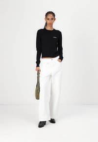 Sort strikket sweater, hvide brede bukser og sorte hælede sko. Modellen holder en grøn taske. Enkel design med minimale accenter.