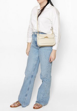 Beige leren schoudertas met een gladde textuur, rechthoekige vorm, gouden accenten en een voorvak. Draagt met lichtblauwe jeans.