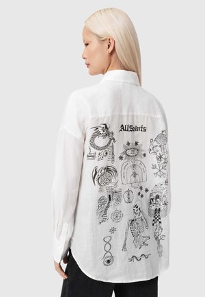 Biała lniana koszula z czarnymi grafikami o różnych wzorach i tekstem na plecach, w tym smokiem, szkieletorem i dekoracyjnymi symbolami.