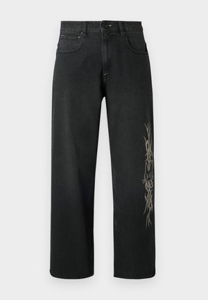 Jean en denim noir avec une coupe ample, cinq poches, une fermeture par bouton et un design graphique prominent sur la jambe gauche.