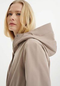 Helle beige wasserdichte Jacke mit großer Kapuze, silbernem Reißverschluss und Druckknöpfen. Glattes Material mit matter Textur, ordentlich taillierte Passform.