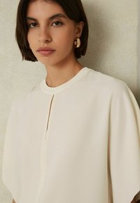 Blusa crema con maniche corte, caratterizzata da un collo rotondo e una fessura frontale. Il tessuto appare liscio e leggero. Orecchini a cerchio dorati visibili.