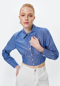Camicia blu a maniche lunghe, con colletto e strisce verticali bianche, tagliata in modo da essere corta, realizzata in un tessuto morbido.