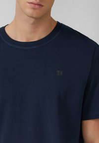 Camiseta de algodón de color azul marino con cuello redondo, que presenta un pequeño logo negro en la parte superior izquierda y una textura suave.