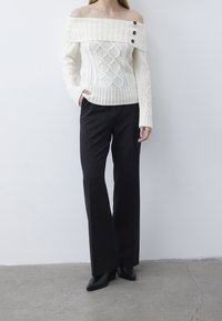 Cremefarbener, schulterfreier Pullover mit Zopfmuster und drei Knöpfen, kombiniert mit schwarzen weit geschnittenen Hosen und spitzen schwarzen Schuhen.