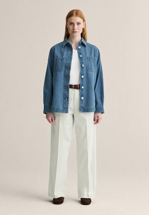 Suorahiuksinen nainen seisoo kasvot eteenpäin, yllään sininen napillinen farkkutakki, valkoinen paita, kermaiset leveälahkeiset housut, ruskea vyö ja ruskeat kengät.