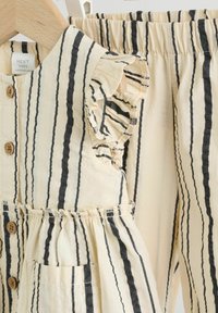 Tenue en coton rayé comprenant un haut boutonné avec des manches à volants et une poche, associé à un pantalon à taille élastique. Les couleurs sont beige et noir.