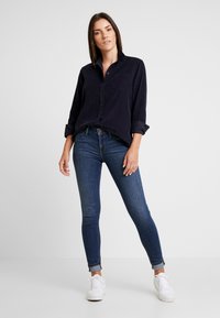 Mörkblå corduroy skjorta med knäppning och bröstficka, i kombination med åtsittande blå denimjeans och vita sneakers. Slät textur och uppkavlade ärmar.