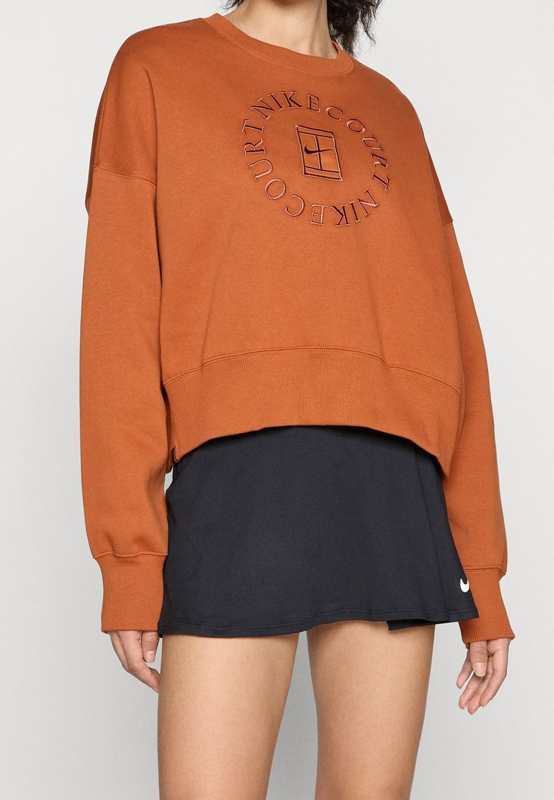 Personne portant un sweat-shirt court orange rouille avec un logo circulaire "Nike Court" et une jupe de tennis noire avec un petit swoosh blanc Nike.