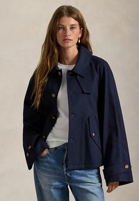 Νεαρή γυναίκα με μακριά μαλλιά φορά navy μπλε κουμπωτό μπουφάν, λευκό t-shirt και μπλε τζιν, στέκεται μπροστά σε απλό φόντο.