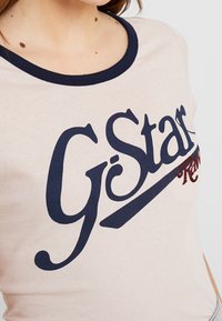 Jasnoróżowy t-shirt z granatowym okrągłym dekoltem. Posiada wyraźne granatowe nadrukowane logo "G-Star" oraz czerwone akcenty szwów.