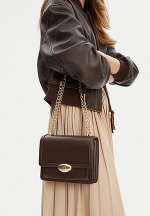Donna che indossa una giacca in pelle marrone scuro e una gonna plissettata beige, con in mano una borsa in pelle marrone scuro con tracolle a catena dorata.