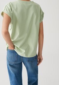 Maglietta verde chiaro con maniche arrotolate, realizzata in tessuto morbido. Abbinata a jeans in denim blu, che mostrano una vestibilità rilassata e dettagli sulla tasca posteriore.