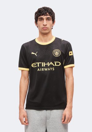 Ung mand iført sort og gul Manchester City fodboldtrøje med Etihad Airways-logo, stående mod en ensfarvet baggrund.