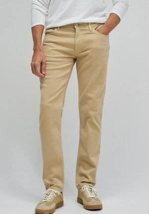 Homme portant un pantalon beige droit, des baskets décontractées beige et blanches, et une chemise blanche à manches longues, une main dans la poche.