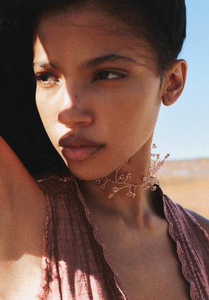 Naszyjnik choker wykonany z delikatnych złotych gałązek z różowymi koralikami. Modelka ma na sobie teksturowaną, jasnoróżową odzież. Naturalne światło tworzy cienie.