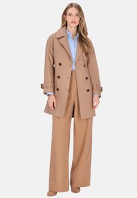 Palton tip „peacoat” maro deschis, cu design cu două rânduri de nasturi mari negri, asortat cu pantaloni largi bej și o cămașă albastră dedesubt.