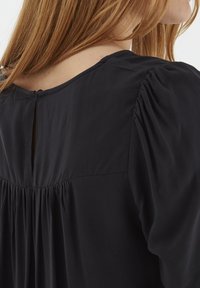 Blusa nera con apertura a occhiello sul retro, dettagli plissé in vita e tessuto morbido e testurizzato con leggeri arricciamenti sulle spalle.