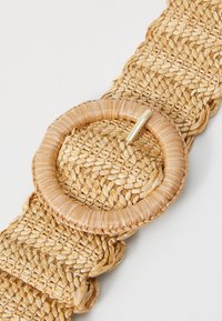 Vevd stråbelte i naturlige toner, med en sirkulær spenne laget av flettet strå og pyntet med en tynn metallpinne for festing.