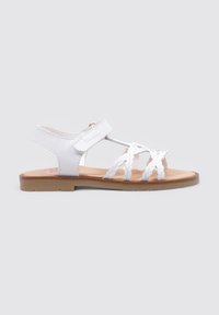 Sandal blanco con tiras trenzadas, correa ajustable en el tobillo y una suela plana con un fondo marrón claro. Textura suave y diseño sencillo.