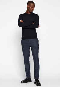 Sötét tengerészkék kötött pulóver, slim-fit kockás nadrág kék árnyalatokban és fekete bőrcipő. A modell karjait összefonva áll, semleges háttér előtt.