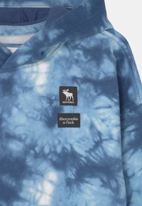 Blauwe tie-dye hoodie met een zachte textuur, voorzien van een centraal logo en een merketiket in contrasterende kleuren.