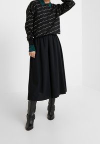 Kobieta w czarnej spódnicy midi, czarnym swetrze z białym napisem, zielonych mankietach i kołnierzyku, oraz czarnych botkach na kostkę z klamrą.